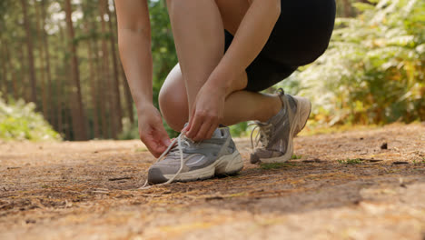 Primer-Plano-De-Una-Mujer-Atándose-Los-Cordones-De-Una-Zapatilla-Deportiva-Antes-De-Hacer-Ejercicio-Corriendo-Por-Una-Pista-A-Través-Del-Bosque,-Filmada-En-Tiempo-Real